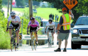 KEEPING RIDERS SAFE as they approach the finish line at New Suncook School on Route 5 in Lovell, Stan Tupaj stops traffic to give cyclists (left to right) Tim Beauchamp, Edward Pond and William Buick a clear path.